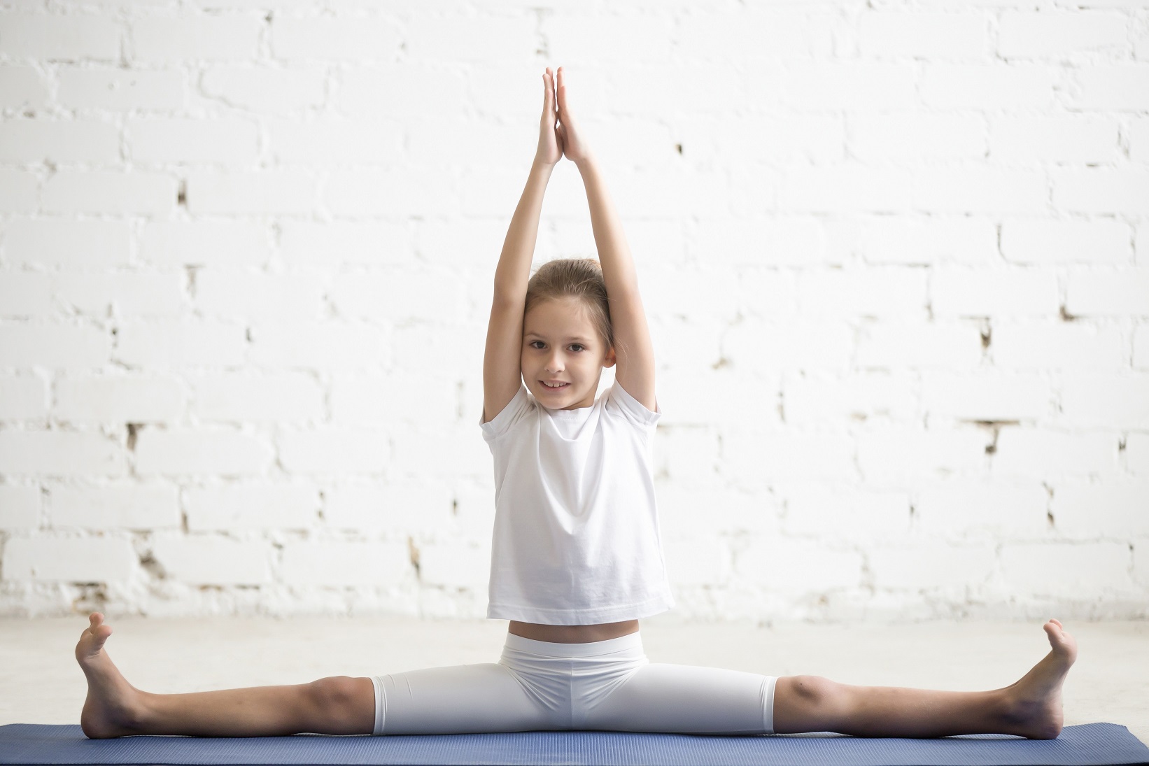 Girl child practicing yoga, doing splits, stretching in Samakonasana exercise, Straight Angle pose, working out wearing sportswear, t-shirt, pants, indoor full length, white studio background
